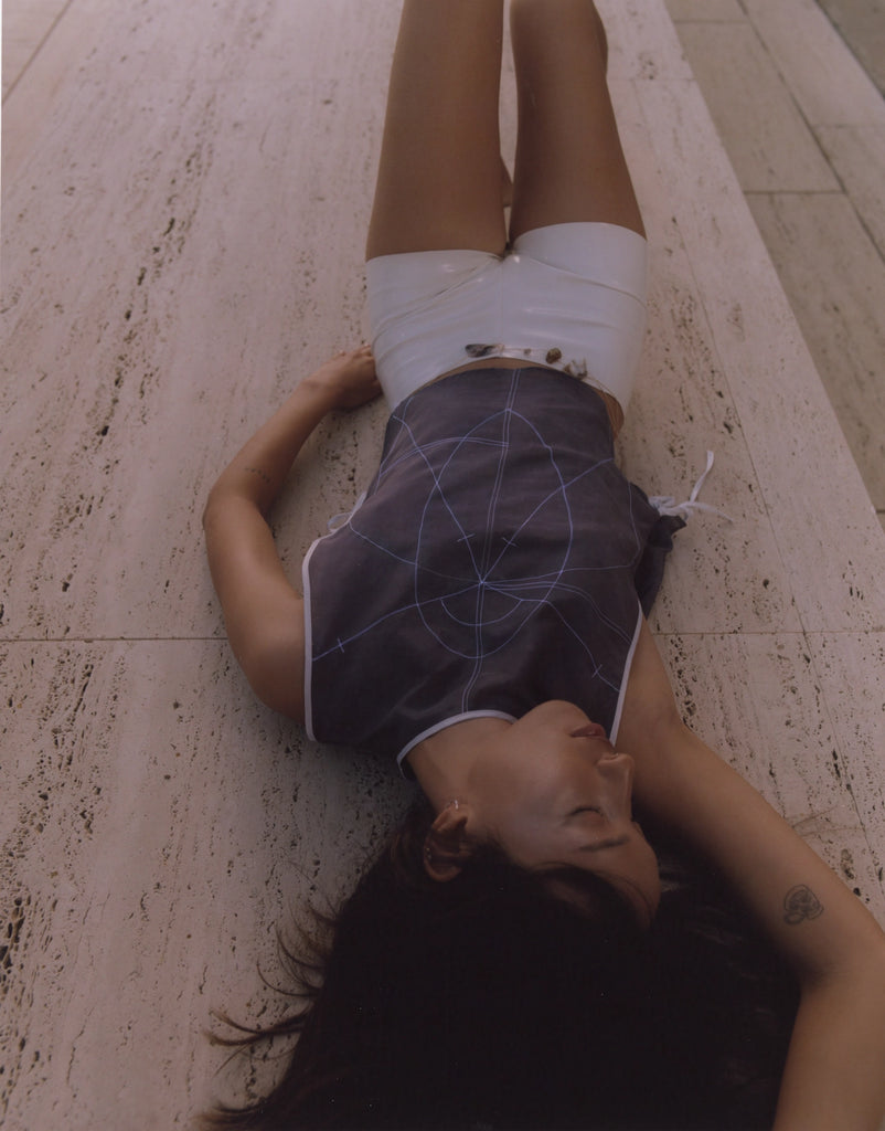 Naaliyah Studios Harmony Top featuring sacred geometry embroidery in white thread, worn by a model lying on travertine stone at the Salk Institute. Indigo silk cotton blend top styled with white shorts in a serene architectural setting